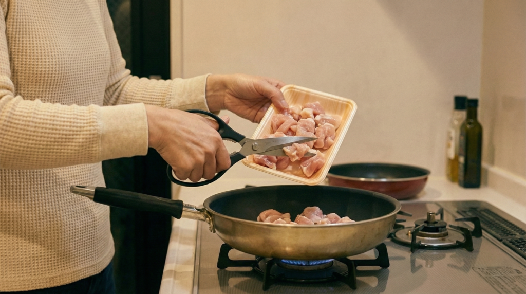 まな板を使わず、キッチンバサミで鶏肉を一口大に切りながら直接フライパンに入れて調理する、効率的で楽しげな様子の女性。